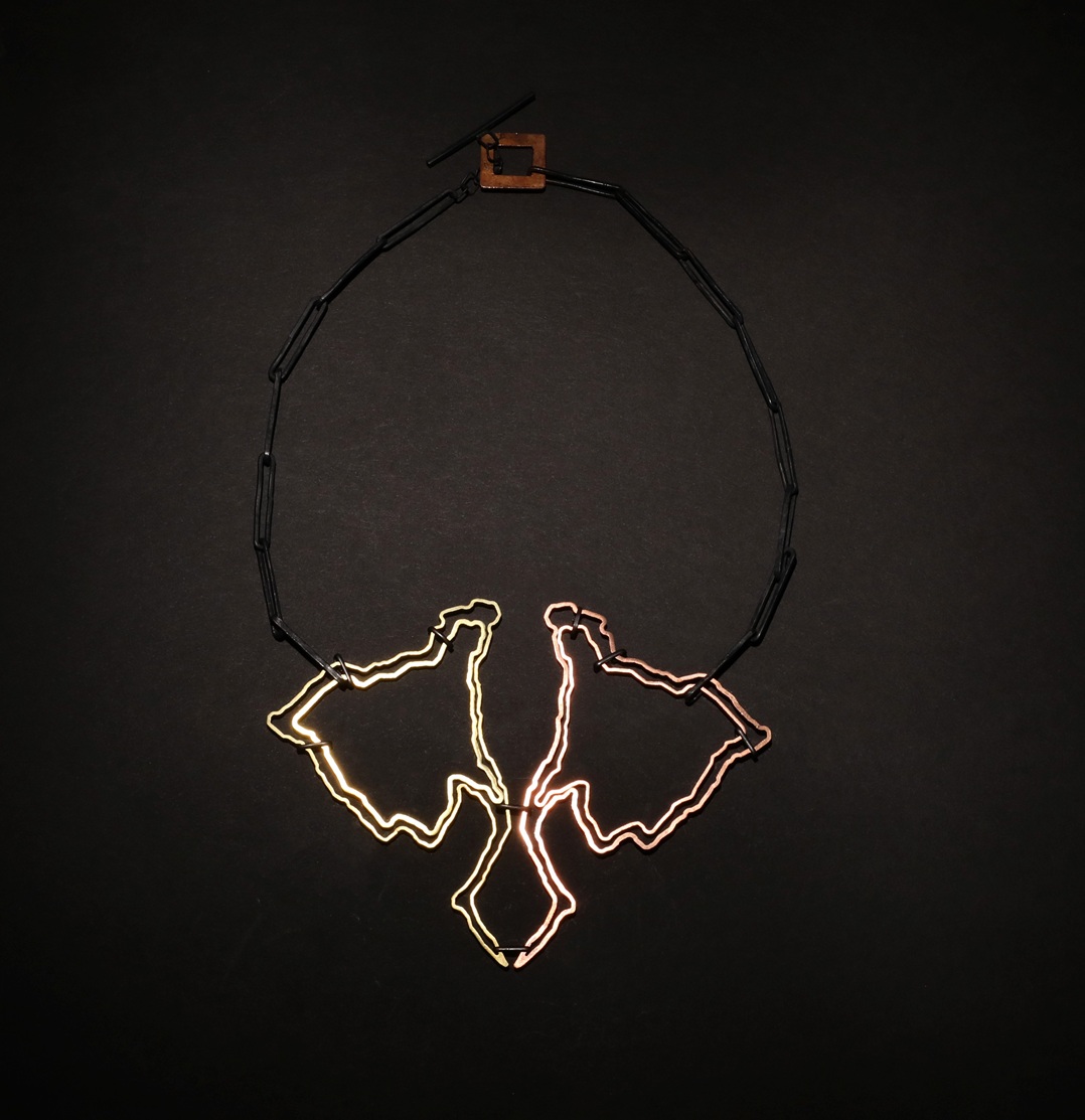 The image shows a single necklace against a black background. The chain is made of dark, rod‑like links forming a loose circle, with a small square wooden clasp at the top. Hanging from the bottom is a large pendant made of thin metal wire shaped into three wavy, abstract, leaf‑like forms. The outlines glow in warm tones—gold on the left, rose‑gold in the center, and copper on the right—meeting at a narrow point before spreading outward like soft, fluttering shapes.