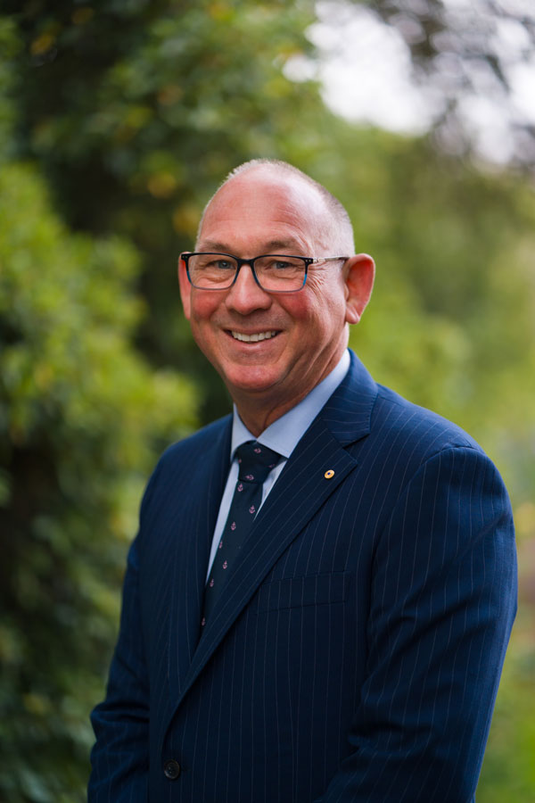 Mal Wise wearing glasses, a navy suit and patterned tie, standing outdoors against a blurred green background.