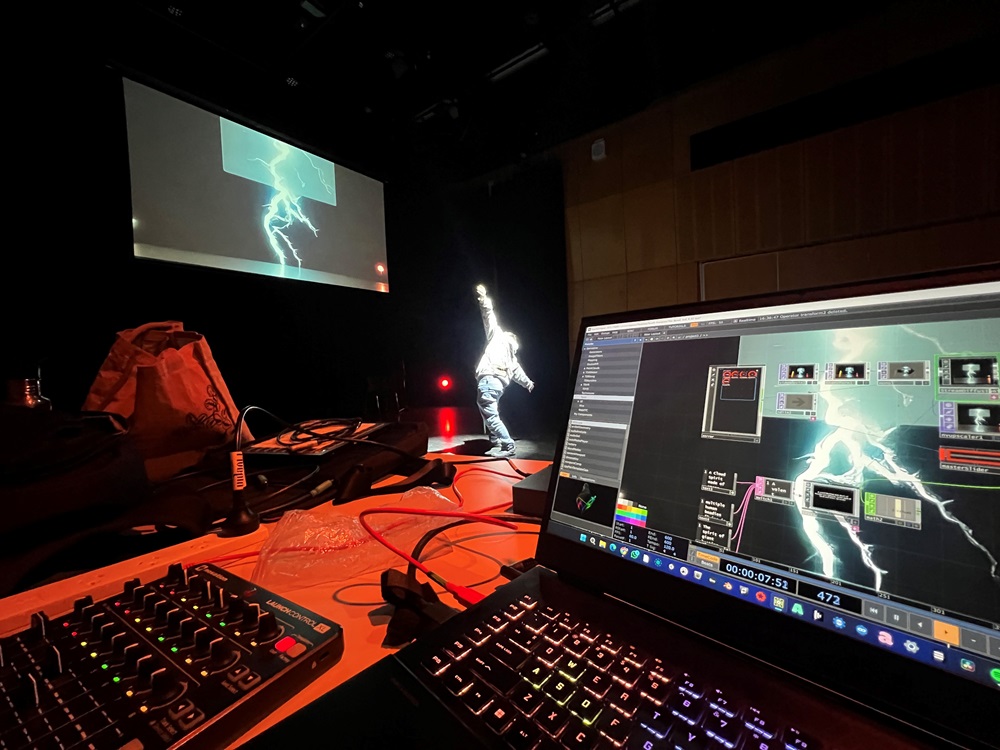 The image shows a multimedia performance using technology. The image shows a workspace with multiple electronic devices, used for audiovisual production or a live performance. There is a large screen displaying an image of lightning, and another smaller projection on the wall showing a similar image. In the centre, there performer with light projections on then creating an effect of being struck by lightning. On the right side of the image, there is a computer with software open that is controlling the visual effects. The foreground shows what appears to be an audio mixer or control surface with sliders and knobs, along with other miscellaneous items like cables strewn across the table.