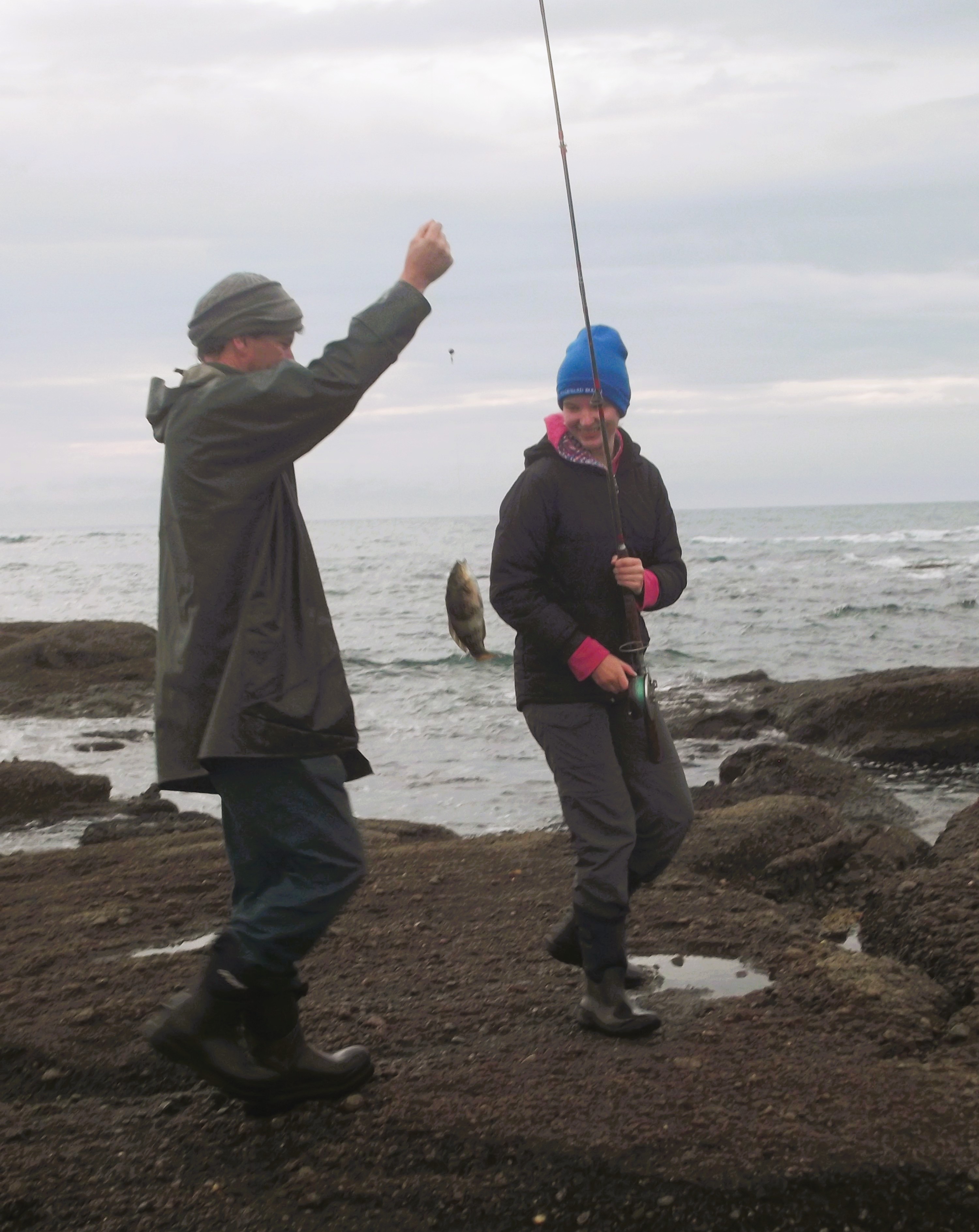 A young woman in a bright blue beanie and black and grey wet-weather gear stands on a pebbled beach, smiling, holding a fishing rod with a fish on the end of the line. A man in dark-coloured wet-weather gear (his face obscured by his raised arm) stands beside her, holding the fishing line.