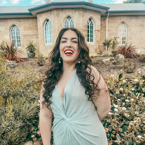 Dana Badcock in a light-colored dress with long, curly hair standing in front of a stone building with arched windows, surrounded by plants and shrubs.