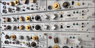 A tall rack of vintage scientific equipment fills the image. It consists of several silver metal panels packed with knobs, switches, dials, sockets, and indicator lights, stacked one above another like drawers in a cabinet.  Many labels read &ldquo;Grass Polygraph&rdquo;, suggesting it is an older physiological recording system. The controls are mostly black, gold, silver, and brown, with small printed markings and measurement scales around them.  The equipment appears well-maintained but dated, giving an industrial, laboratory feel. A dark curtain hangs along the left side, while the silver panels dominate the frame under bright, even lighting.