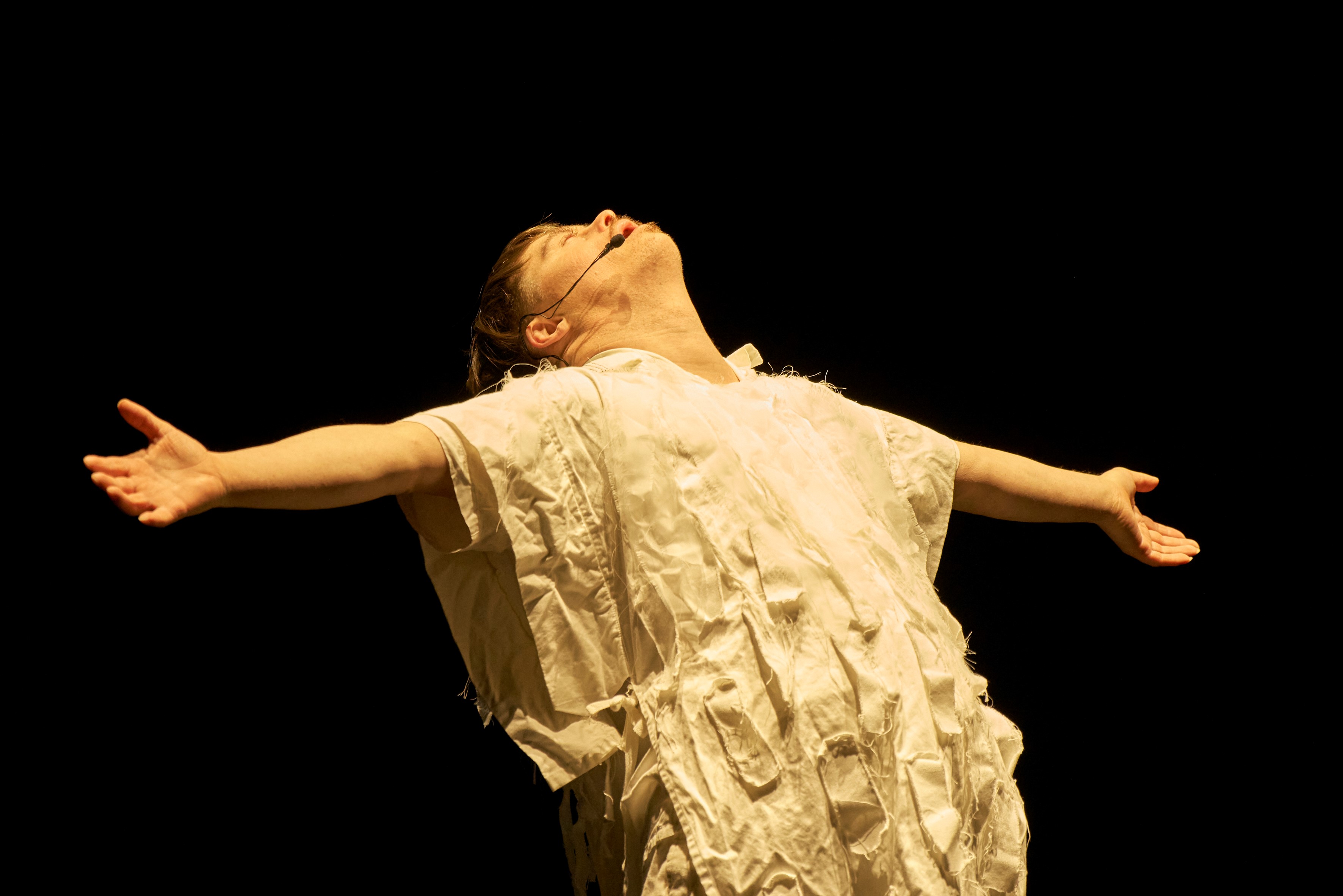 The image portrays a person (Luke John Campbell) standing on a stage, seemingly engaged in a dramatic performance. Their head tilts back, eyes closed, and arms stretch wide, suggesting an expression of release or embracing the moment. The individual wears a textured, layered garment resembling a dress or costume with ruffled elements. The background is entirely black, emphasizing the figure as if illuminated by a spotlight.