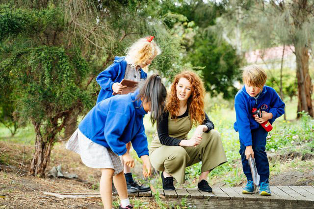 A female teacher kneeling on a wooden path with three children in school uniforms, exploring nature in a bush setting