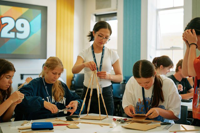 A group of students is seated around a table in a brightly lit classroom, working on a hands-on project with wooden sticks, cardboard, and other craft materials.