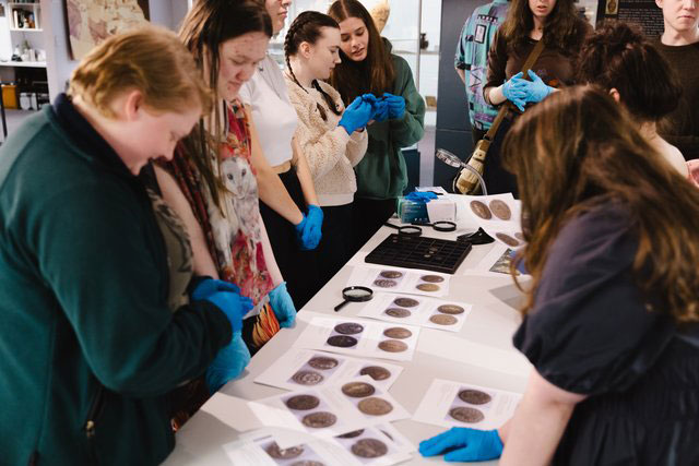 Students wearing blue gloves examine printed sheets of ancient coins on a table with magnifying glasses and trays, inside a museum setting.
