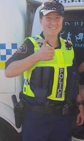 A female Tasmania Police officer smiles and gives a thumbs-up gesture. She is wearing a blue police uniform and cap with a yellow high-vis vest.