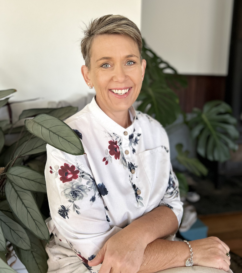 Kristi Chapple sitting indoors beside large leafy plants, wearing a light floral-patterned shirt.