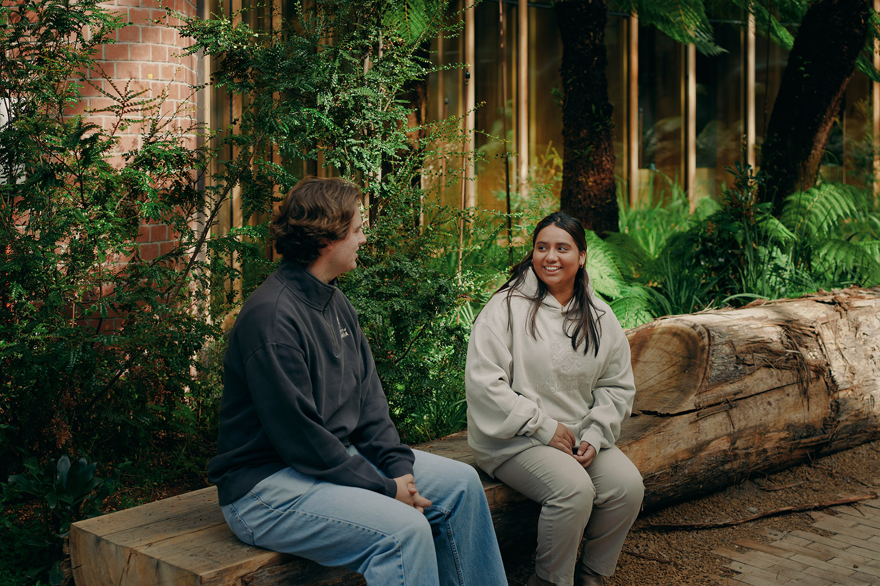 Two students seated on a large timber bench in an indoor garden space, surrounded by greenery and natural wood features in the new Forestry building.