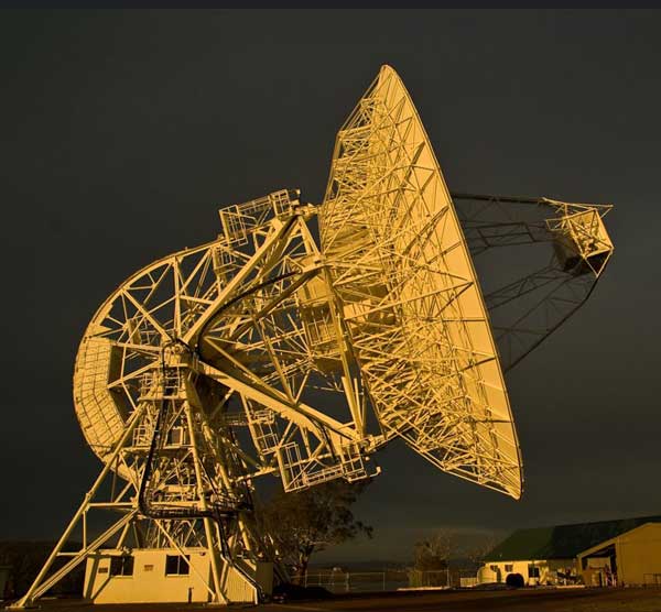 Radio telescope dish at sunset, angled skyward with buildings and trees in the background.