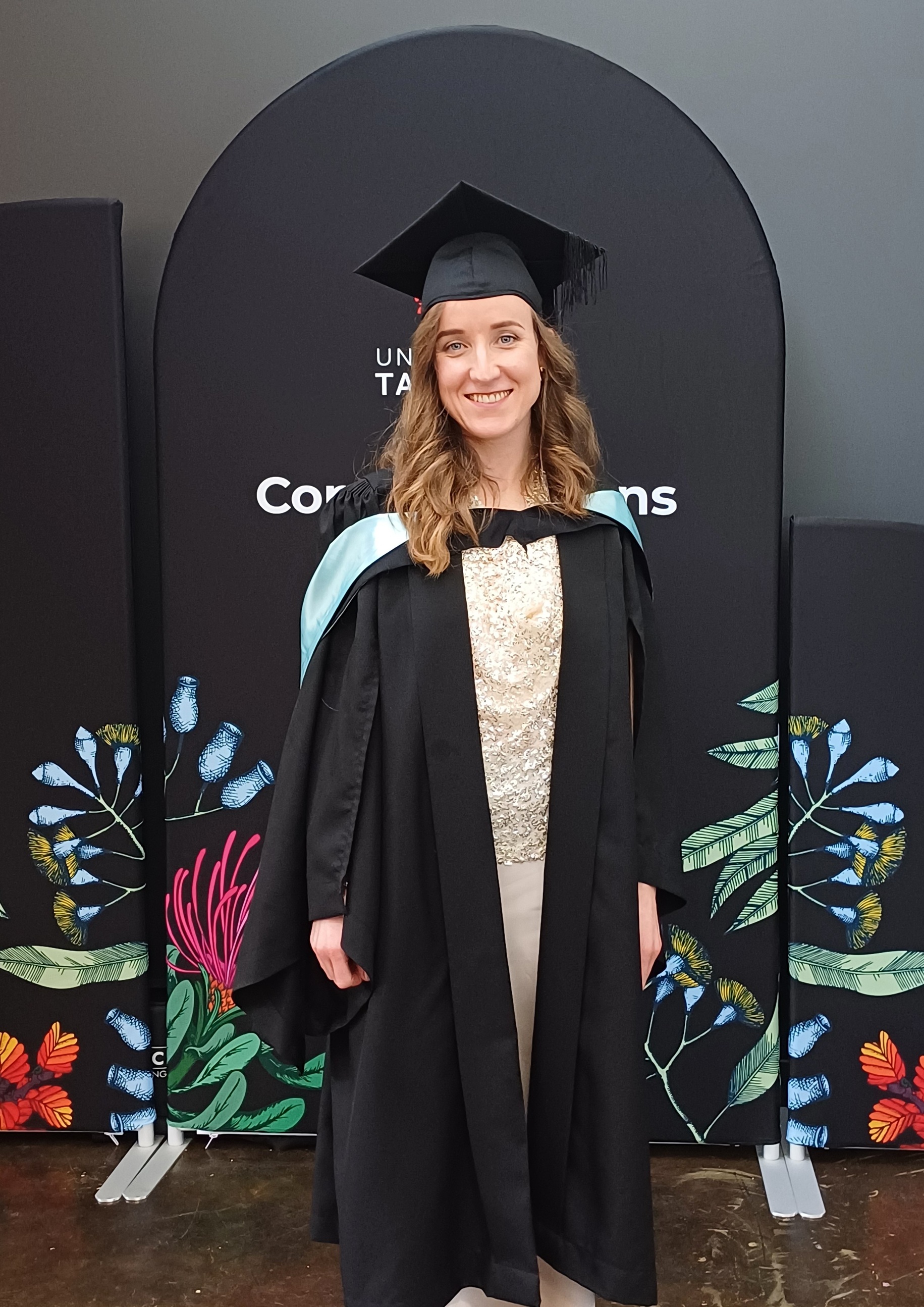 A young woman with shoulder-length, medium brown hair smiles at the camera, wearing a black academic gown and black mortar board, with the light blue-lined hood of an education graduate.