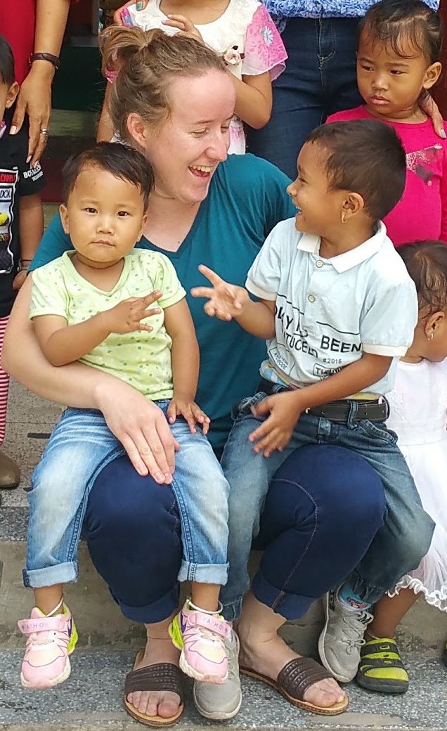 A caucasian woman with light brown hair in a ponytail, wearing blue jeans and a turquoise t-shirt, laughs as she holds a young Nepalese child on each knee. The child on the right is looking back at her and laughing.