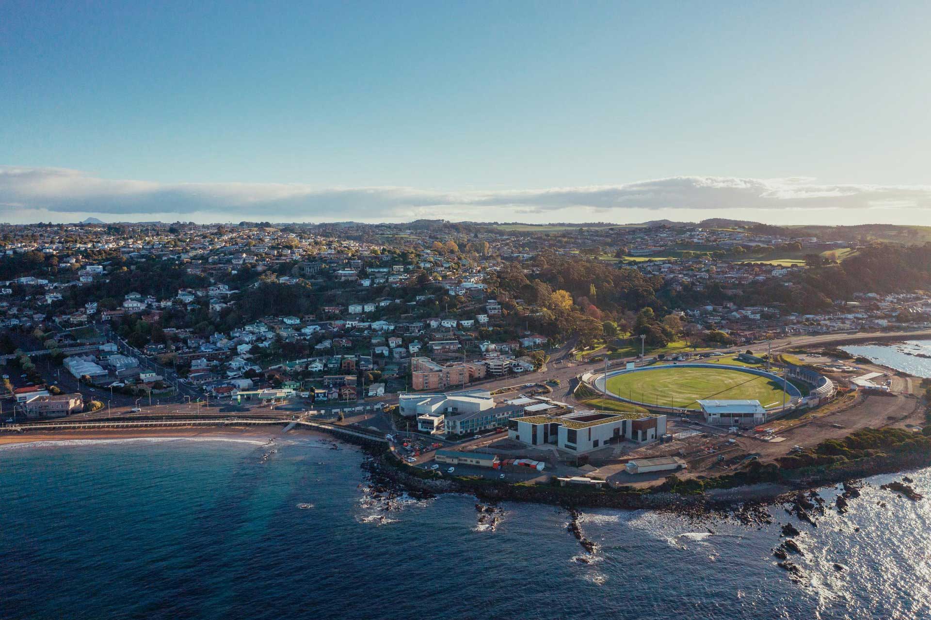 Aerial view of West Park a large sports field near the rocky shoreline, surrounded by densely packed houses extending into hillside terrain.