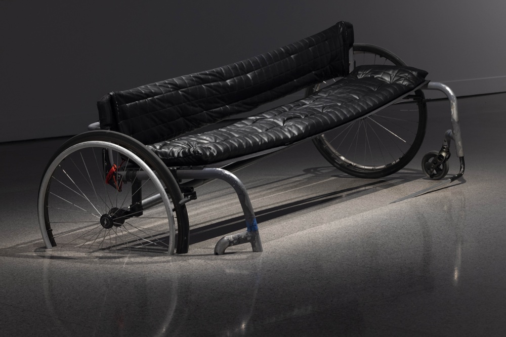 This image shows a bench with a sculptural shape that clearly resembles a mobility aid. The seat and backrest are padded in black, providing a comfortable, soft, quilted feel that stands out against the harder materials of the frame.  The structure of the bench is made from metal tubing, giving it a modern, industrial look. On each side, there are large wheels similar to those found on wheelchairs.  The bench is placed on a smooth, painted grey floor.