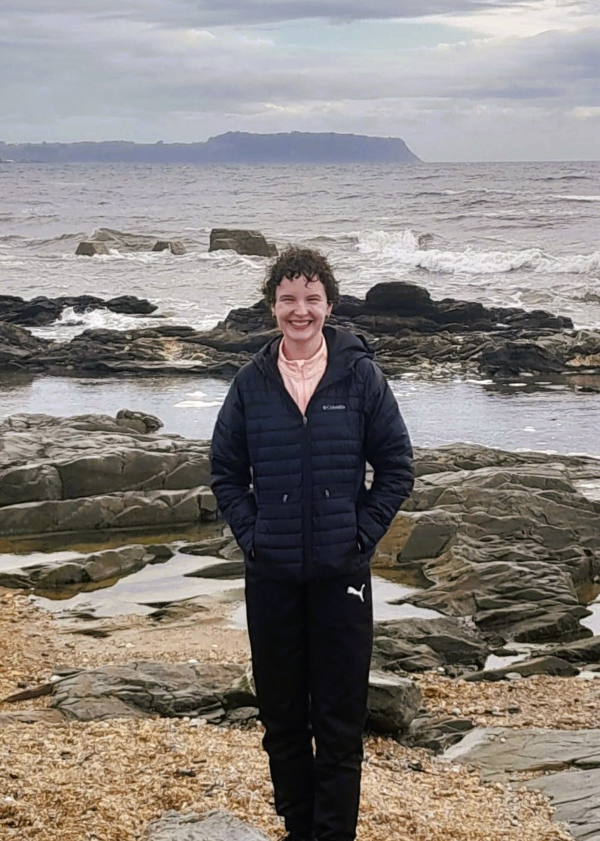 Trinity Shires stands on the shore of a rocky, pebbled Tasmanian beach, overcast skies and grey choppy water in the background. She has short, curly, dark brown hair, fair skin, and is smiling at the camera. She is wearing black trackpants and a black puffer jacket.