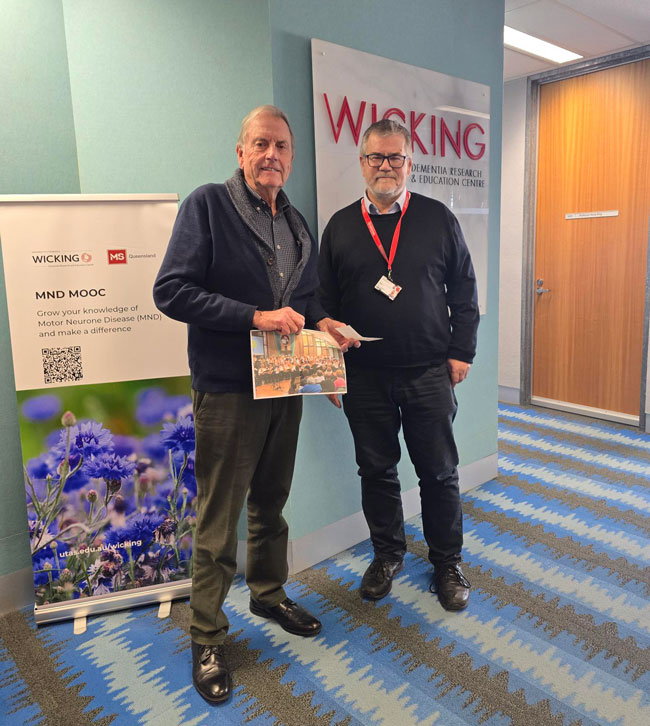 Lauchlan and James standing beside a Wicking Centre poster promoting MND awareness and education
