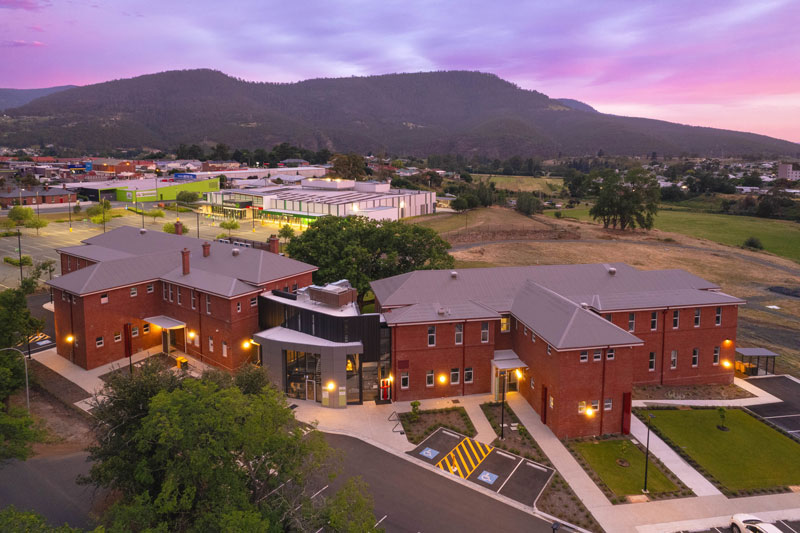 Aerial view of a large red‑brick building complex at sunset, surrounded by green fields, nearby facilities, and distant hills under a purple sky.