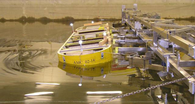 Yellow model ship secured to a metal testing rig in an indoor water tank, fitted with sensors and labeled “AMC 02-14,” with gentle ripples on the water surface.